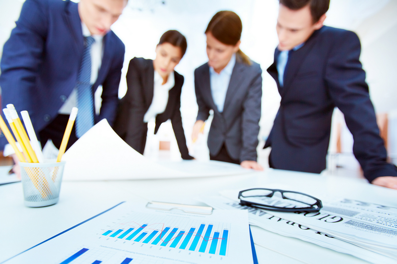 Four business professionals discuss documents on a desk with charts, pencils, and glasses in focus.